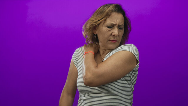 Woman in striped shirt holds shoulder against purple studio wall with closed eyes in pain; discomfort.