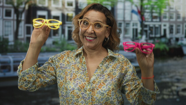 Woman holds colorful eyeglasses in each hand by canal front of building in amsterdam; joy playfulness choice.