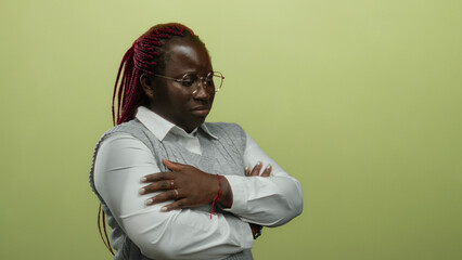 African american woman with braided hair in glasses and gray vest stands with arms crossed over isolated yellow background, conveying confidence and contemplation.