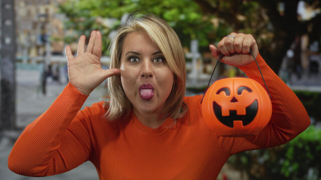 Woman wearing orange sweater holding jack o lantern bucket sticking tongue out and waving hand on street; playfulness.