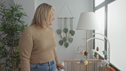 Woman smiling with hand on crib rail by window at mobile in building; maternal nurturing bonding serenity.