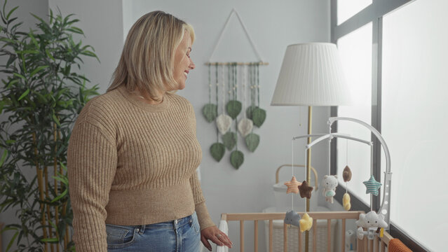 Woman smiling with hand on crib rail by window at mobile in building; maternal nurturing bonding serenity.