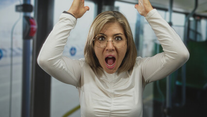 Obraz premium Woman in white shirt and glasses opens mouth and raises arms inside crowded city bus during commute near window seat; surprise reaction.