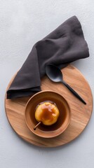 Overhead of a half-caramelized baked pear in a minimalist bowl on a wooden serving board with a black spoon.