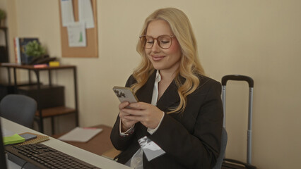 Woman working in an office using smartphone wearing glasses and a black suit sitting indoors with a suitcase in the background suggesting a business environment