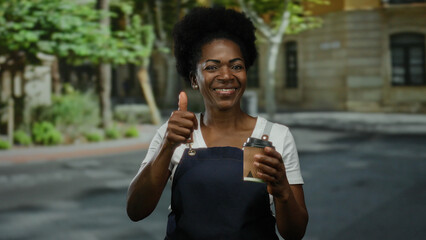Woman smiling holds coffee cup outdoors on city street wearing apron expressing positivity and approval with thumbs up gesture exuding friendly and approachable vibe with urban backdrop