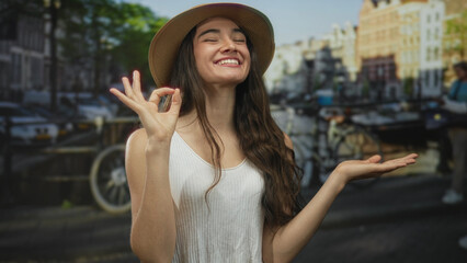 Woman wearing straw hat and white tank top making ok sign with hands on street canal bridge under sunlight; approval optimism.