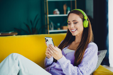 Young woman with green headphones sits on a yellow sofa at home using a smartphone enjoying music and relaxation in a bright modern living room