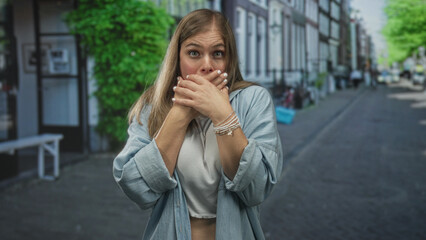 Fototapeta premium Young blonde woman gently covers mouth with both hands amid city buildings on a paved street; surprise.