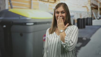 Fototapeta premium Young blonde woman with hand covering mouth in front of large recycling container on busy street; surprise.