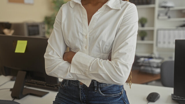 African american woman in white blouse and jeans with arms crossed at office desk inside a building, computer monitors and files visible; confidence.