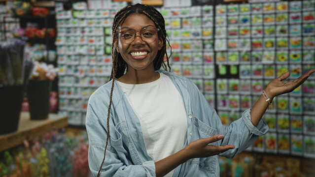 African american woman smiling and gesturing with open hand in a flower shop wearing glasses and denim shirt braided hair visible; joy.