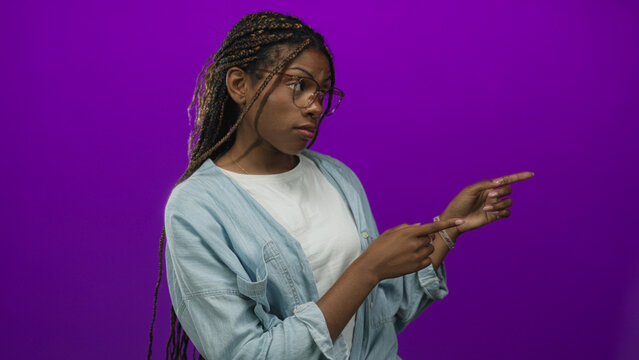 Woman pointing with index finger and glancing sideways while wearing glasses and denim shirt in a vibrant purple studio setting; doubt. - Powered by Adobe