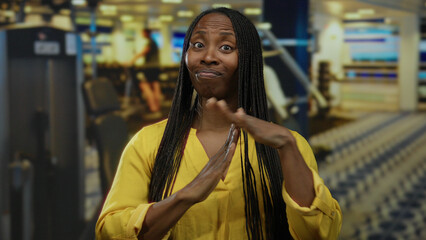 Woman with braided hair making time gesture wearing yellow shirt in gym setting with blurred fitness equipment in background.