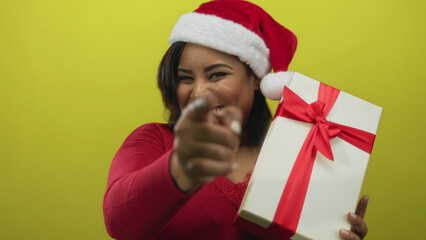 Woman wearing santa hat holds gift against yellow wall celebrating christmas with joy and cheer pointing happily.