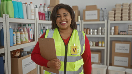 Young woman volunteer in a reflective vest holds clipboard inside donation center surrounded by boxes and products, showcasing dedication to charity work in a supportive environment.