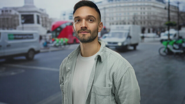 Young man with beard stands on urban street with traffic in background, wearing casual shirt and white t-shirt, while city buildings frame the image vividly.