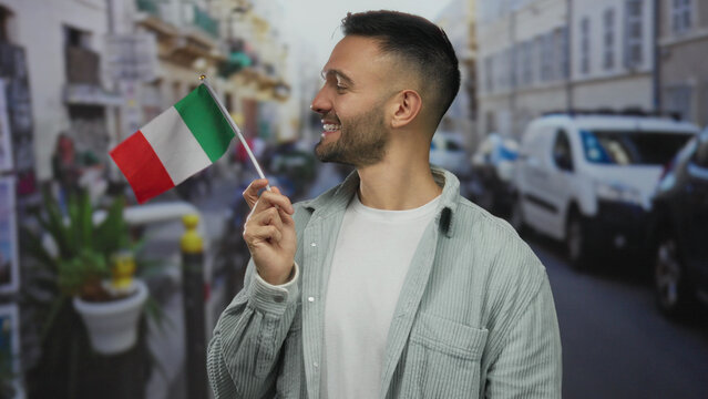 Fototapeta Young man smiling and holding italian flag in an urban street setting with cars and people visible in the background, conveying a vibrant city atmosphere.