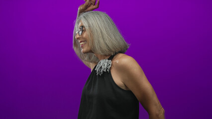 Woman dancing with raised bare arm and smiling face in purple studio, grey hair, glasses, black halter top; joy celebration.
