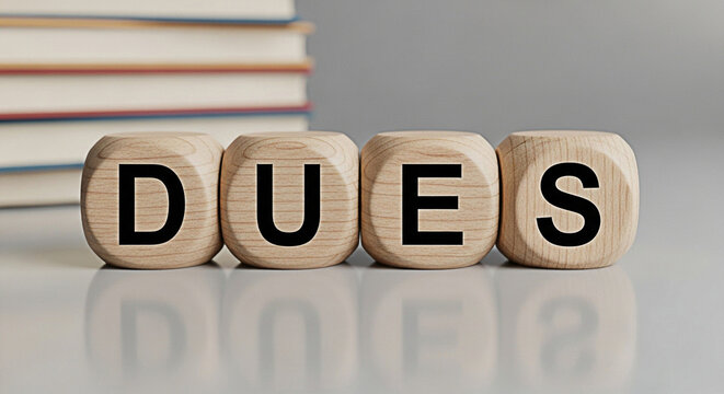 Wooden blocks spelling "dues" with books in the background represent financial obligations and membership fees in an educational setting