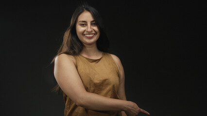 Fototapeta premium Woman pointing finger with bare hand and exposed arm in black studio wearing brown tanktop and smiling; playful confidence.