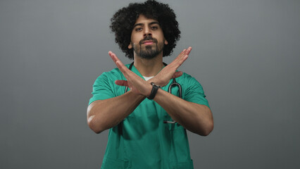 Man in green scrubs wearing a stethoscope crossing forearms in a grey studio setting; denial caution.