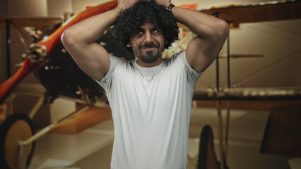 Man with curly hair and beard, hands on head beside vintage biplane in museum exhibit, wearing white tshirt and showing biceps; nostalgia amusement.