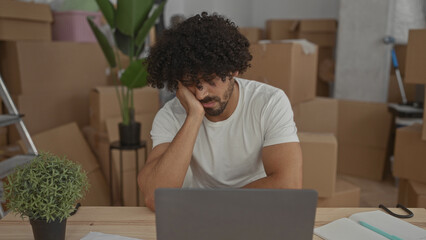 Man with hand on cheek in building filled with cardboard boxes using laptop and notepad; fatigue unpacking.