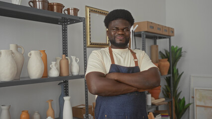 Young african american man standing with arms crossed, wearing denim apron, smiling beside pottery vases on metal shelves in a studio; pride.