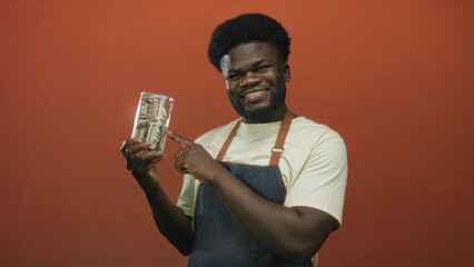 Man points finger to clear glass jar of rolled cash and dollar bills on orange studio wall;...