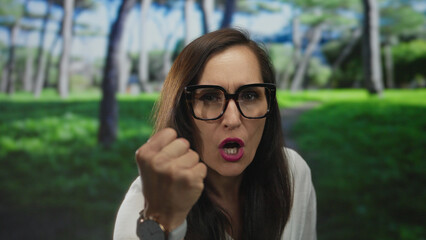 Woman with glasses gesturing outdoors in vibrant green park surrounded by tall trees on a sunny day...