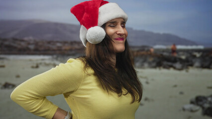 Young hispanic woman holds lower back at seaside amid rocky shoreline under overcast sky; backache discomfort.