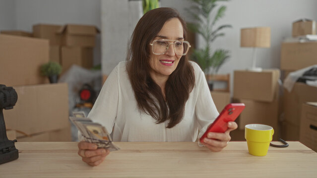 Woman using smartphone in new home surrounded by unpacked boxes holding us dollars, showing satisfaction in an indoor room environment.