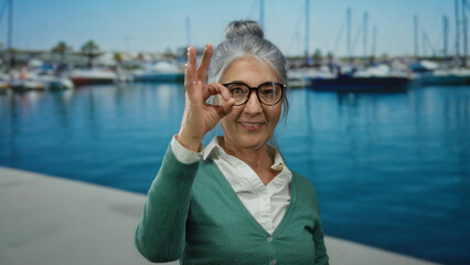 Senior woman smiling by the seaside with boats at port, making okay gesture, wearing glasses, grey hair in an outdoor scenic view, perfect for capturing nautical vibes.