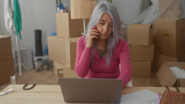 Senior woman with grey hair talking on phone while working on laptop in her new home surrounded by cardboard boxes, highlighting a modern, independent lifestyle transition.