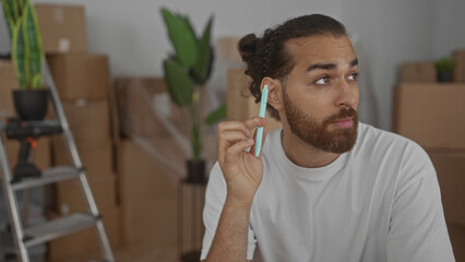 Man holding mint pen near ear and looking sideways while seated among packed moving boxes in building; pensive planning.