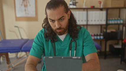Man doctor in green scrubs with stethoscope reading medical tablet at desk in clinic; duty focus patient care.