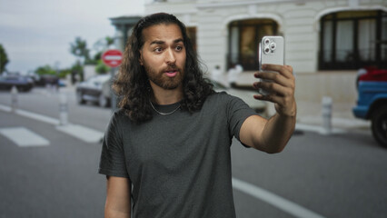 Man holding smartphone for selfie on city street with extended arm and open mouth; curiosity sharing.