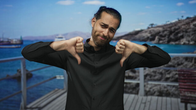 Man showing thumbs down with both hands on a seaside street pier near wooden boardwalk and docked boats; disapproval.