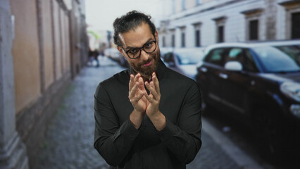 Man with glasses rubbing fingers together in black shirt on cobblestone street; scheming slyness...