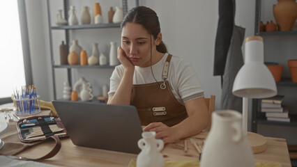 Young hispanic artisan woman wearing brown apron with hand on cheek typing on laptop in studio;...
