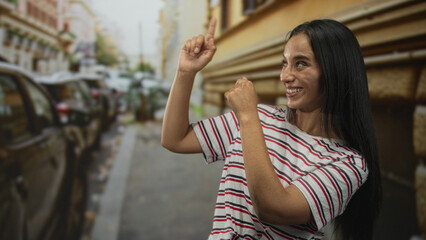 Woman points fingers upward on a city street while smiling and leaning on a building wall, wearing a striped t shirt; joy discovery curiosity.