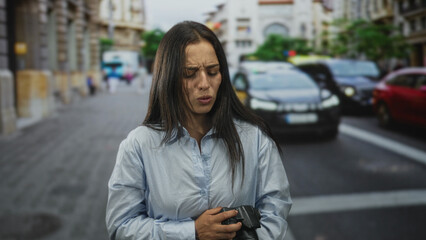 Hispanic woman clutching a camera at a crosswalk on a busy city street, wincing with a pained...