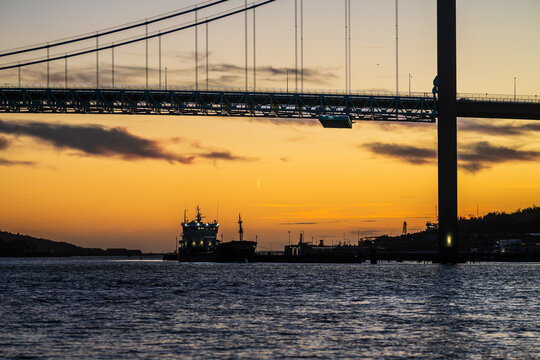 Cargo ship sailing under bridge at twilight with orange sky. - Powered by Adobe