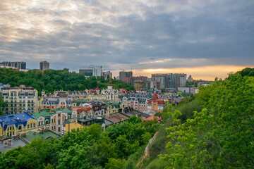 Obraz premium Kyiv cityscape showcasing the colourful houses of Vozdvyzhenka district nestled amongst lush green trees, with modern buildings and a bright horizon under a cloudy sky at sunset