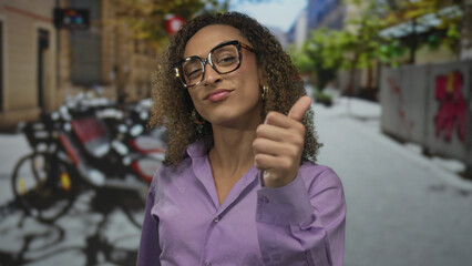 Transgender woman in purple blouse and stylish glasses gives a thumbs up gesture on a street; approval confidence positivity optimism.