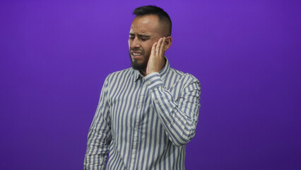 Young hispanic man in a striped shirt holding hand to ear and wincing in a purple studio with toothache; pain.