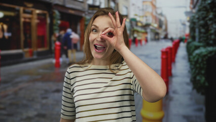 Woman wearing striped shirt makes ok sign around eye on urban street with red bollards and blurred pedestrians; curiosity exploration discovery wonder.
