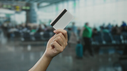 Man holding credit card at airport terminal with blurred background of travelers waiting.