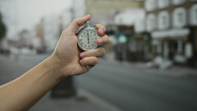 Hand holding a stopwatch in an urban street setting with blurred town buildings, symbolizing time management and precision by a man in an outdoor city environment.
